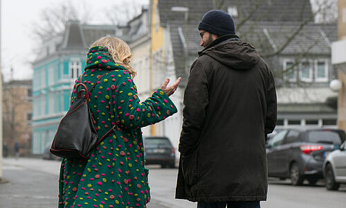 Eine Frau und ein Mann von hinten beim Gang auf einem Bürgersteig. Beide tragen hüftlange Mäntel, auf dem Asphalt sind kleine Pfützen.
