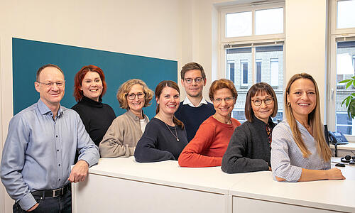 Ein Gruppenfoto des Teams der Geschäftsstelle der Brücke Flensburg in den Geschäftsräumen an einem weißen Tresen. Alle lehnen sich auf den Tresen und lächeln.